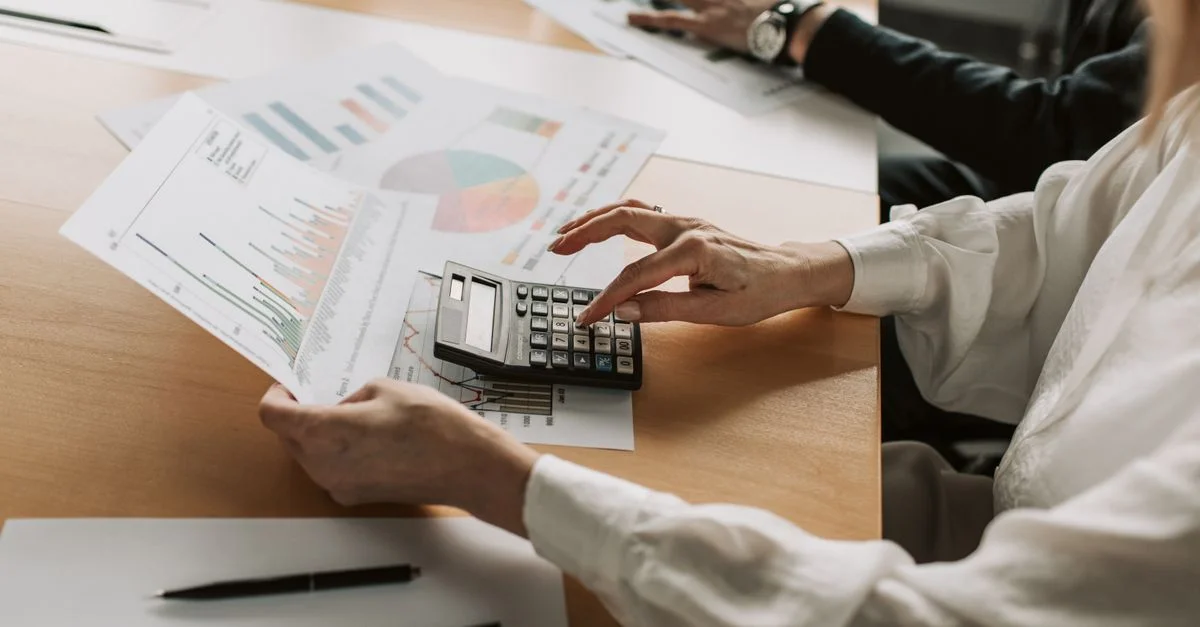 Manos realizando cálculos financieros con gráficos y una calculadora en una mesa de reuniones.