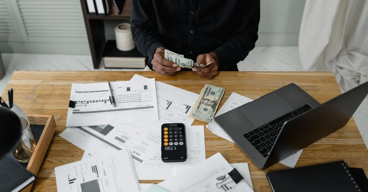 Una persona cuenta billetes de dólar en un escritorio con una laptop, papeles y una calculadora.