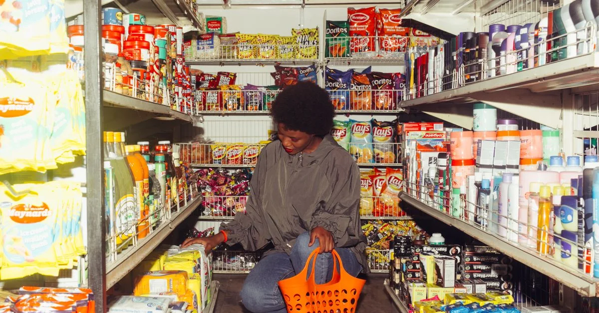 Mujer afroamericana seleccionando productos en una tienda de conveniencia con una cesta naranja.