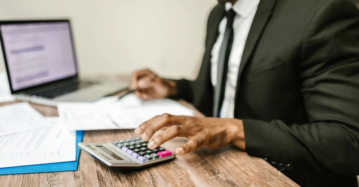 Un empresario con traje negro usando una calculadora en un escritorio con documentos financieros y un portátil.