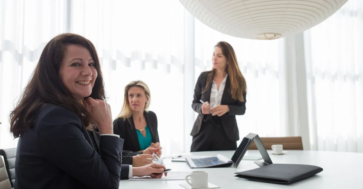 Tres mujeres vestidas formalmente sonriendo y discutiendo negocios durante una reunión.
