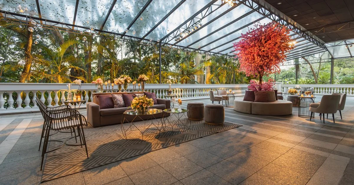 Configuración lujosa de terraza al aire libre con sofás, mesas y decoración floral bajo una cubierta de vidrio.