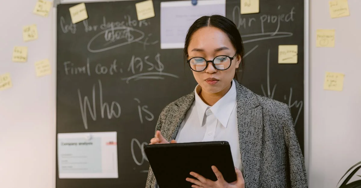 Mujer asiática en una oficina usando una tableta digital, usando gafas, de pie frente a una pizarra.
