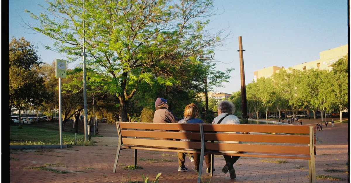 Tres personas sentadas en un banco del parque bajo árboles, disfrutando de un entorno urbano tranquilo.