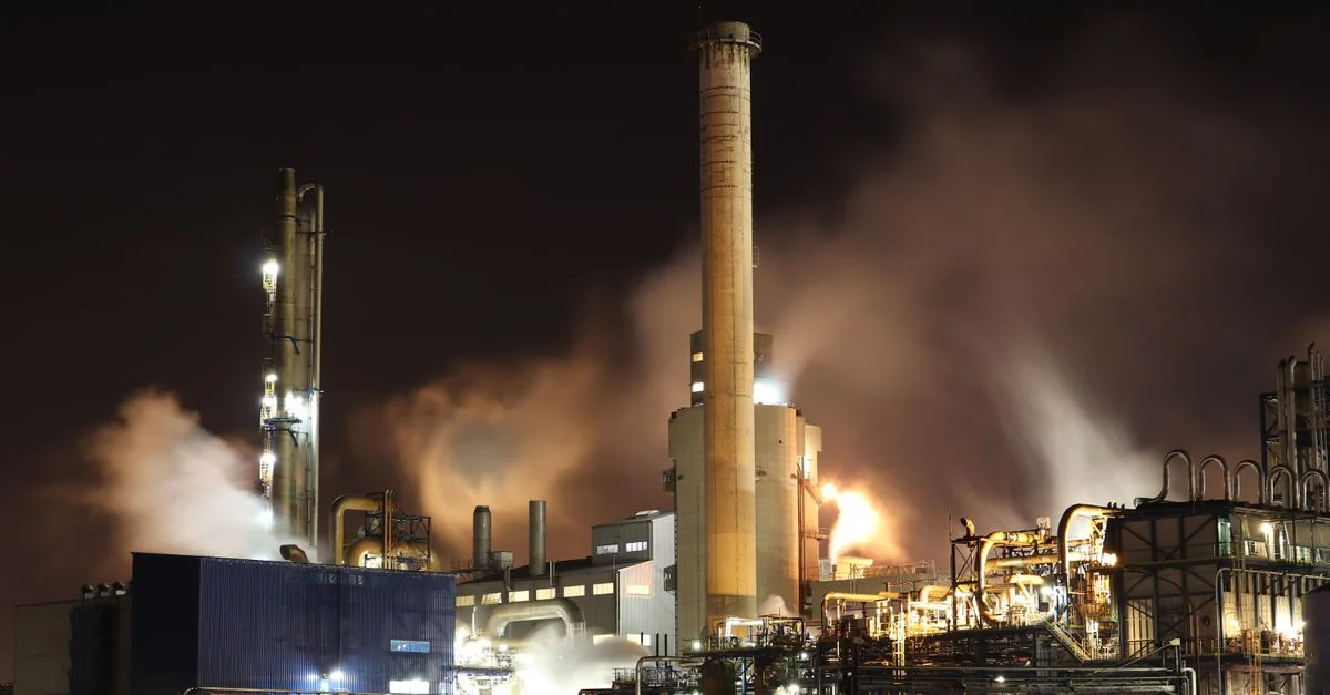 Una vista nocturna de una fábrica industrial con chimeneas que emiten humo, destacando la producción de energía y la contaminación.
