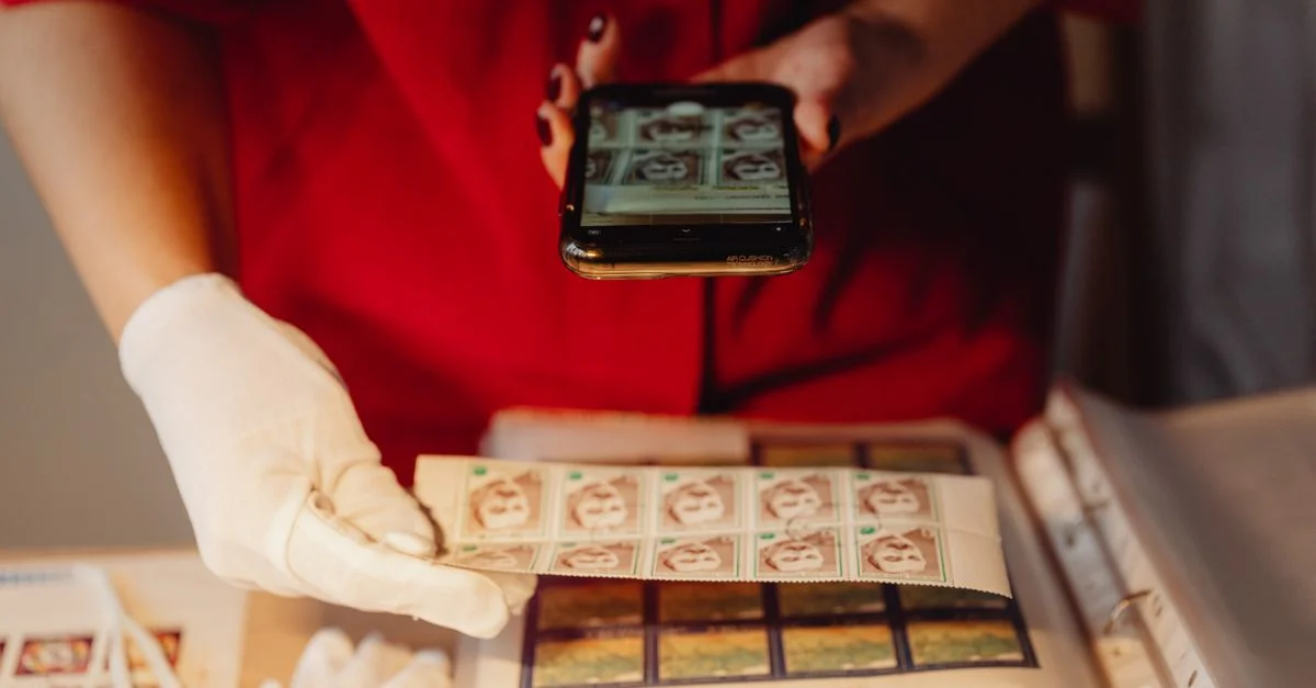 Una persona con guantes fotografía una colección de sellos utilizando un teléfono inteligente para archivo digital.